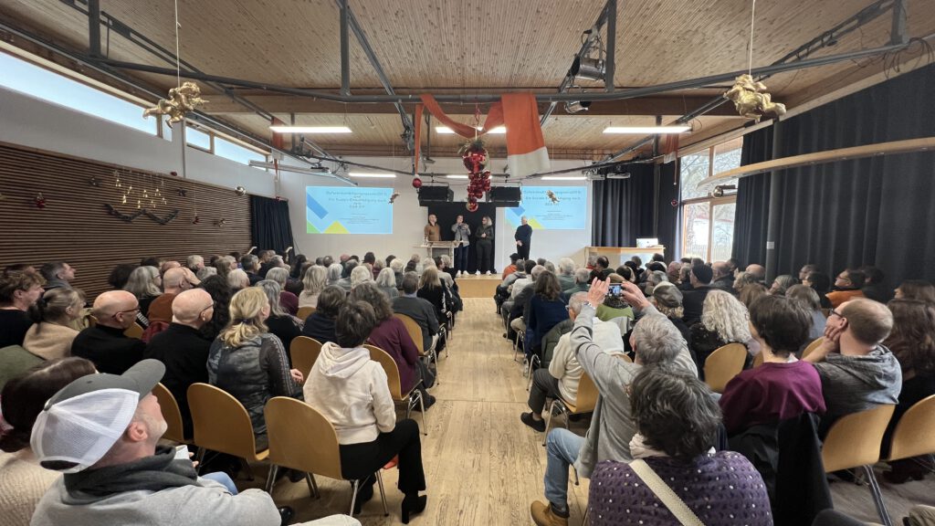 Von hinten: viele Zuschauende, auf der Bühne stehen Stephan Straßer, Can Sipahi, Cornelia von Pappenheim und Christian Ebmeyer. Auch sind zwei Leinwände links und rechts zu sehen. Der Saal ist weihnachtlich geschmückt. 