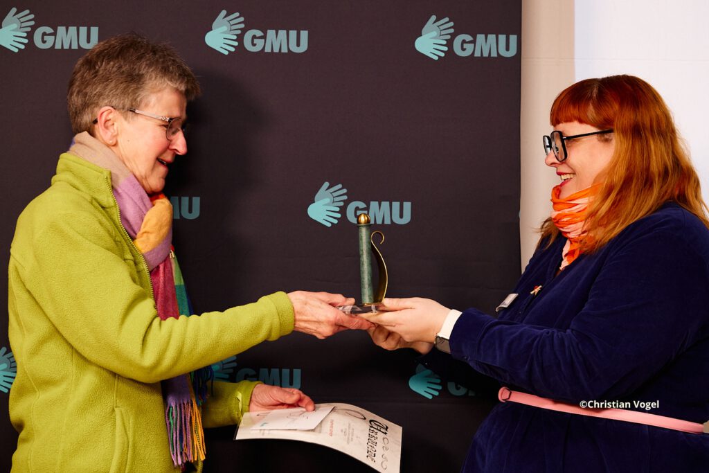 Rosemarie und Lina stehen sich gegenüber und halten gemeinsam eine Auszeichnung in den Händen. Beide lächeln sich an. Im Hintergrund ist ein dunkler Banner mit GMU-Logos zu sehen.