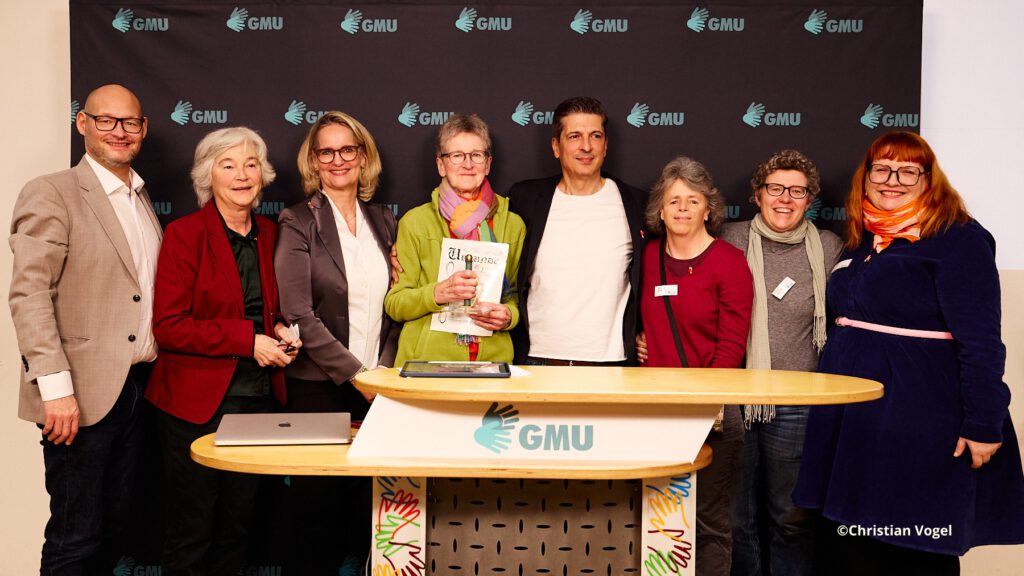 Im Hintergrund ist ein dunkler Banner mit GMU-Logos zu sehen.
8 Personen stehen und lächeln.