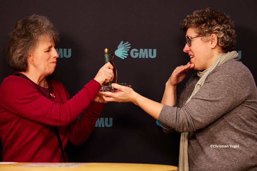 Martina bekommt eine Auszeichnung von Doris.
Im Hintergrund ist ein dunkler Banner mit GMU-Logos zu sehen.