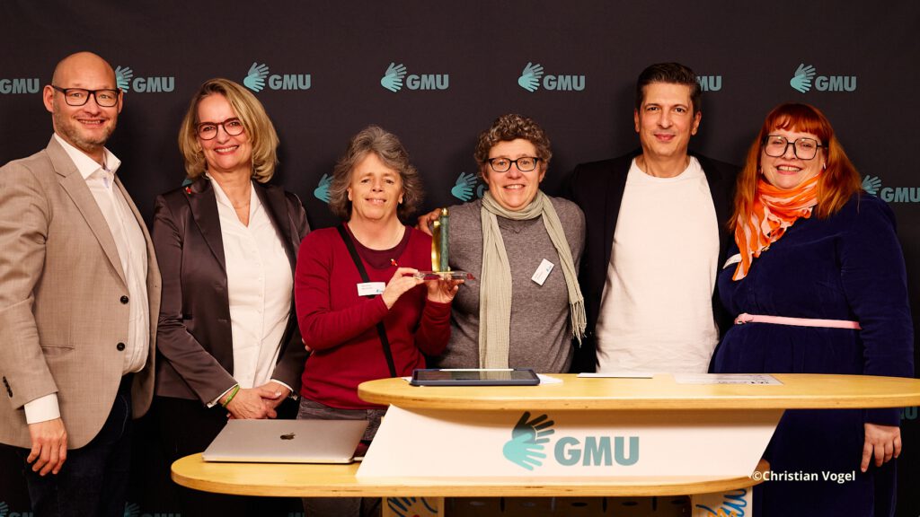 Im Hintergrund ist ein dunkler Banner mit GMU-Logos zu sehen.
6 Personen stehen und lächeln.