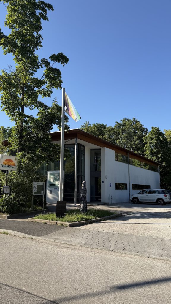 Außenansicht eines modernen, zweigeschossigen Gebäudes mit weißer Fassade und großen Glasflächen an einem sonnigen Tag. Vor dem Eingang weht an einem Fahnenmast die Disability-Pride-Flagge. Das Gebäude ist von hohen Bäumen umgeben, links steht ein Schild mit der Aufschrift „Locanda Tramonto“. Rechts neben dem Gebäude ist ein geparktes weißes Auto zu sehen. Der Himmel ist wolkenlos und blau.